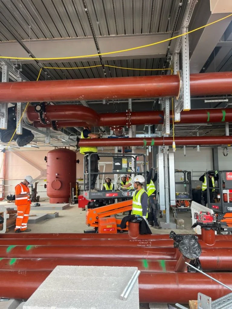 Workers in high-visibility clothing install large red pipes inside a building under construction.